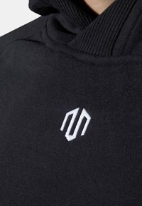Tejido de sudadera negra con una textura suave. Presenta un cuello acanalado y un logo bordado en blanco situado en el pecho.