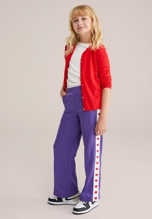 Ung tjej står med händerna i fickorna på lila byxor, klädd i en röd cardigan över en vit topp och svart-vita sneakers.
