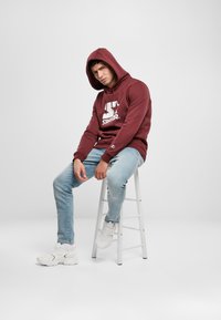 Felpa bordeaux con logo bianco, jeans e sneakers bianchi. Il modello è seduto su uno sgabello bianco, mostrando un design di streetwear casual.