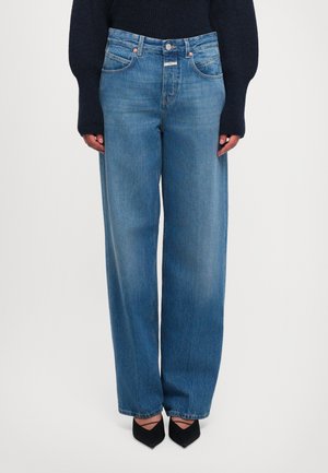 Femme portant un jean bleu taille haute à larges jambes, des talons noirs à bout pointu avec des brides, et un pull noir à manches longues, se tenant droite.