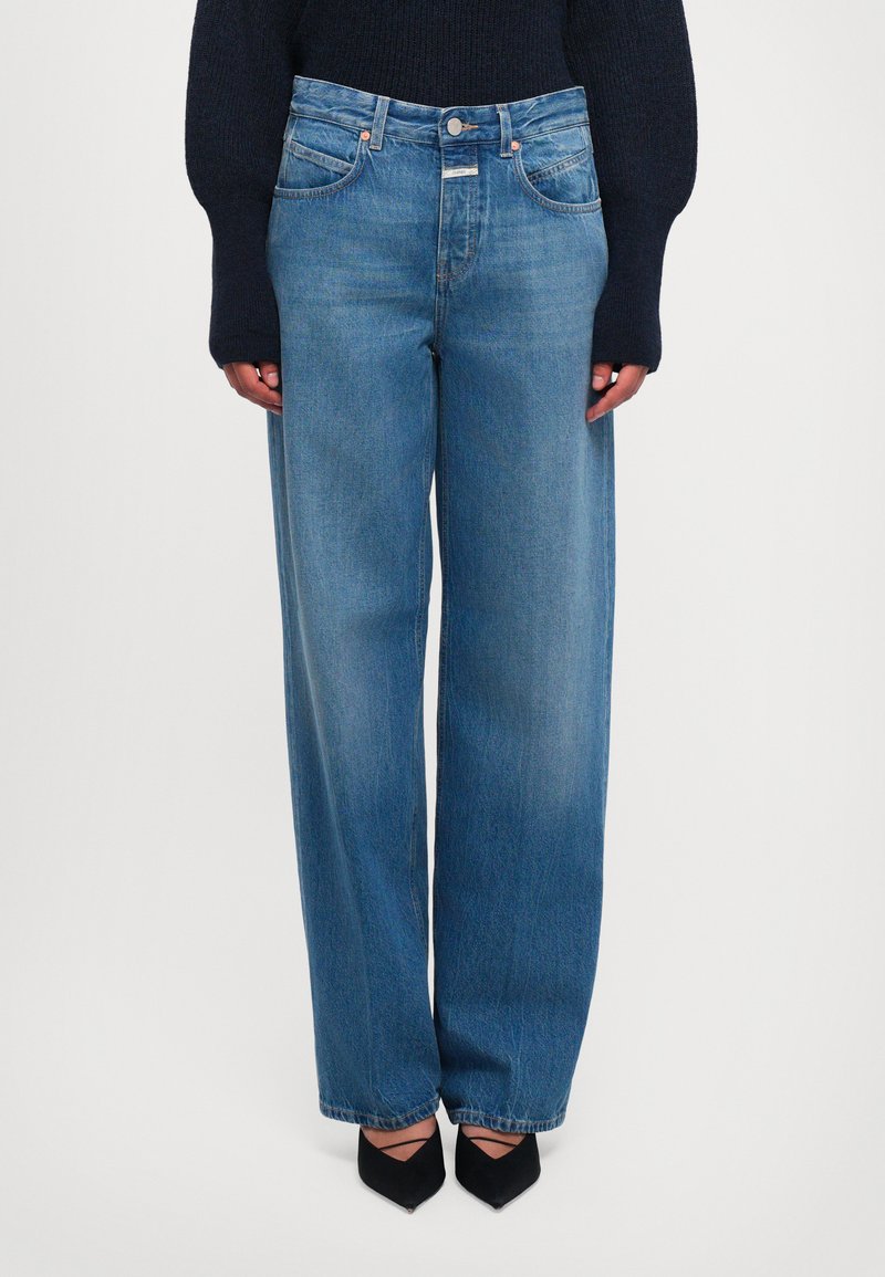 Vrouw in hoge taille wijduitlopende blauwe jeans, zwarte puntige hakken met banden, en een zwarte lange mouwen trui, rechtop staand.