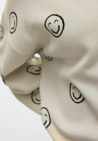 Sweter w kolorze kremowym z czarnymi haftowanymi uśmiechami rozproszonymi po tkaninie, charakteryzujący się miękką teksturą i luźnym krojem.