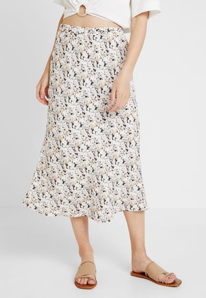 Kvinde iført en hvid midi nederdel med blomsterprint kombineret med beige flade sandaler med åben tå og en hvid top med en ringdetalje foran.