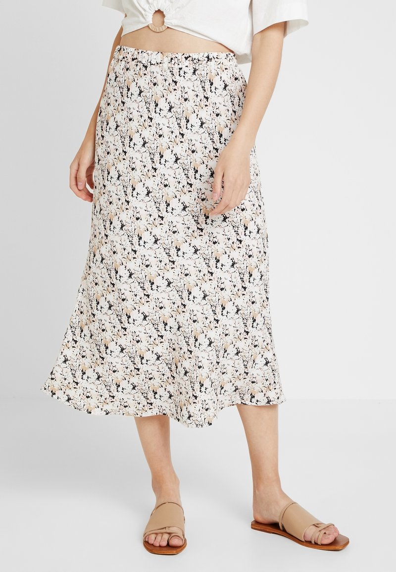 Femme portant une jupe midi blanche à motif floral, associée à des sandales plates beiges à bout ouvert et un haut blanc avec un détail anneau à l'avant.