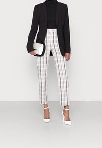 Černý blazer s úzkým střihem, spojený s kostkovanými kalhotami v bílé, hnědé a béžové barvě. Bílé vysoké podpatky a strukturovaná bílá kabelka.