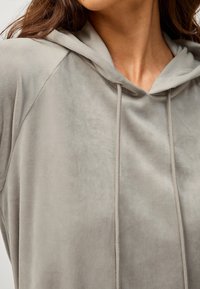 Šedá fleecová mikina s kapucí, která má měkkou texturu, obsahuje šňůrku na stažení a raglánové rukávy pro uvolněný střih.