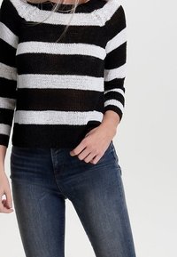 Femme portant un pull en maille rayé noir et blanc à manches trois-quarts et un jean slim bleu foncé, debout devant un fond uni.