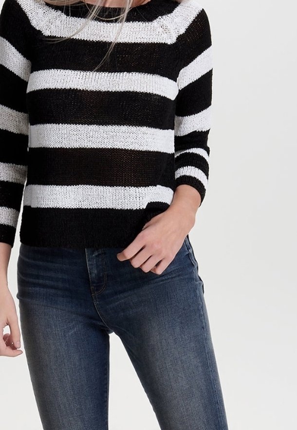 Femme portant un pull en maille rayé noir et blanc à manches trois-quarts et un jean slim bleu foncé, debout devant un fond uni.
