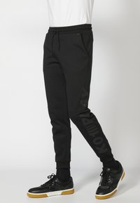 Sorte sporty joggerbukser med snoretræk i taljen, slim fit, sidelommer og en mat logo trykt lodret på venstre ben.