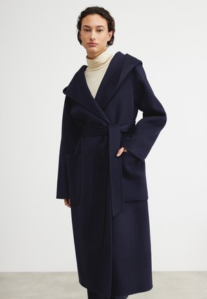 Cappotto in lana navy con ampio colletto, vita cintata e grandi tasche frontali. Il tessuto testurizzato e le maniche lunghe creano una silhouette strutturata.