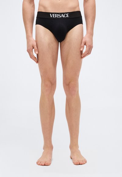 Modèle masculin portant un slip noir Versace avec ceinture logo, debout pieds nus devant un fond clair uni.