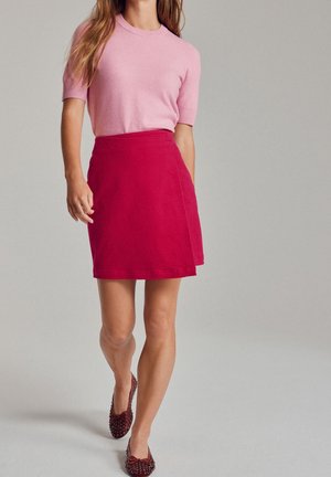 Mujer vistiendo una blusa de manga corta rosa, falda roja y zapatos planos burdeos con tachuelas, caminando frente a un fondo gris liso.