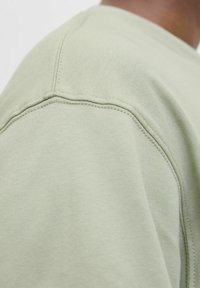 Hanorac de culoare verde deschis, cu o textură netedă, detalii vizibile de cusături și un design cu guler rotund. Vedere detaliată a zonei umărului.