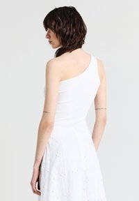 Top bianco a costine con una spalla, dalla forma aderente. Modella che indossa una gonna bianca con ricami floreali. Sfondo neutro.