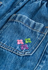 Denimkangas, jossa on vaaleansininen sävy; siinä on tasku, jossa on vaaleanpunaisia, vihreitä ja violetinvärisiä kukka-ompeluksia; näkyvät ompelutiedot.