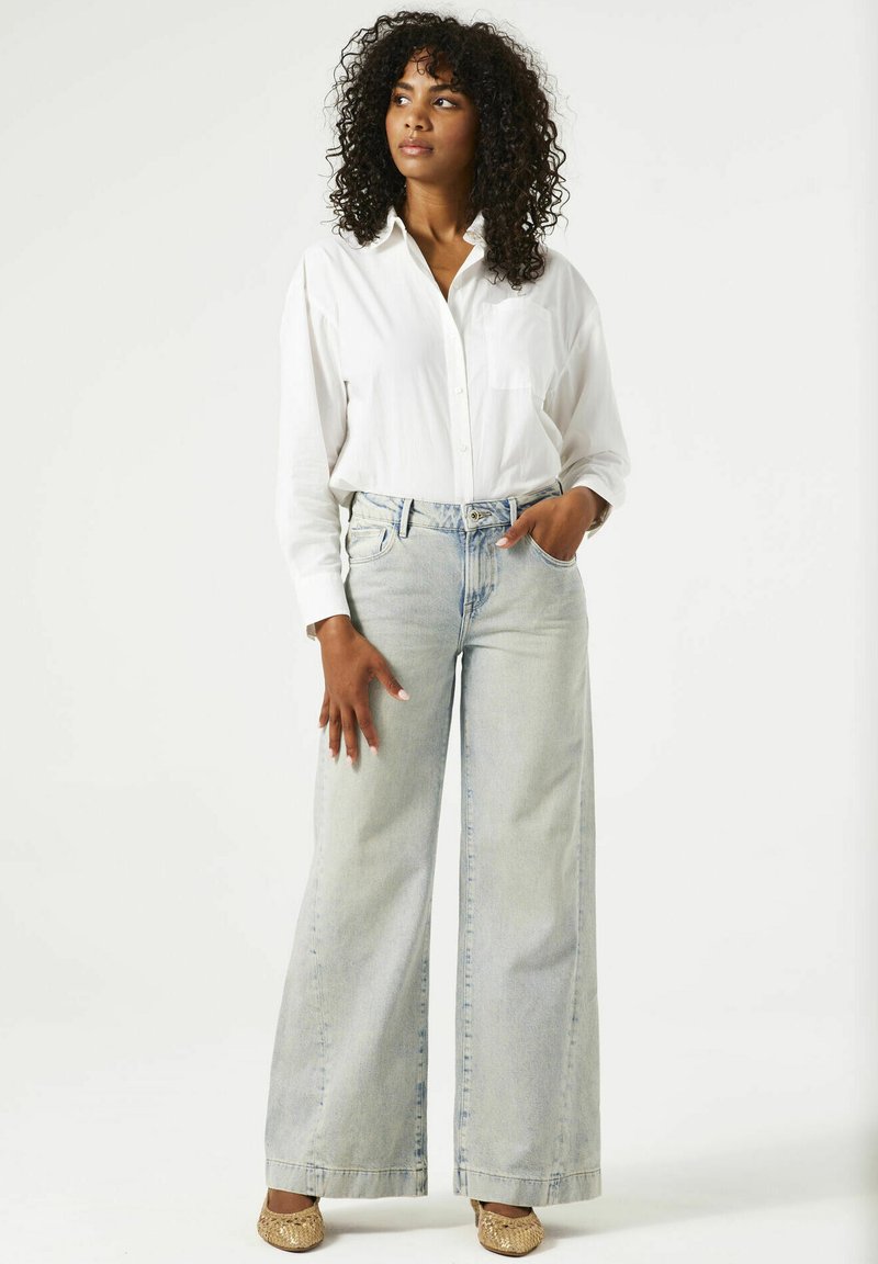 Camisa blanca de manga larga con botones, jeans de pierna ancha azul claro y bailarinas doradas. Telas suaves con un ajuste relajado y detalles de bolsillo en el frente.