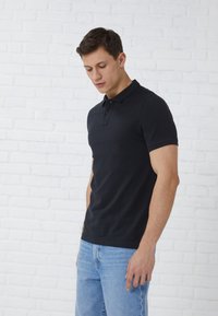 Polo shirt noir en coton avec une patte de boutonnage à trois boutons, associé à un jean en denim bleu clair, vu de côté contre un mur en briques blanches.