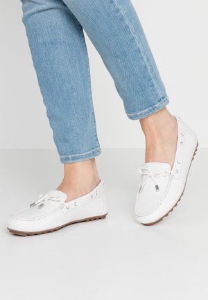 Füße, die hellblaue Jeans und weiße Leder-Mokassins mit Gummisohlen tragen, gezeigt vor einem einfarbigen hellen Hintergrund.