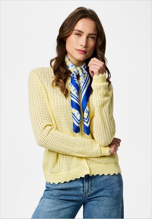 Kvinde med langt brunt hår iført en gul strikket cardigan, blåt blomsteret tørklæde og blå jeans, poserer med den ene hånd nær ansigtet.