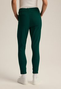 Zielone dopasowane spodnie z elastycznym pasem, wykonane z elastycznego materiału. Posiadają tylne kieszenie i gładką fakturę. Połączone z białymi sneakersami.