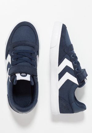 Granatowe dziecięce trampki z białymi paskami w kształcie szewronu, białą podeszwą, sznurowadłami i paskiem na rzep na białym tle.