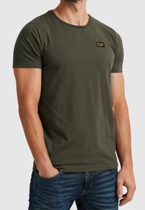 Man wearing a fitted olive green short-sleeve t-shirt with a small black and yellow chest logo, paired with blue jeans.