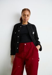 Schwarze Strickjacke mit silbernen Knöpfen, kombiniert mit dunkelroten, hochgeschnittenen Hosen mit Taschen und einer seitlichen Schlaufe. Schwarzes Oberteil darunter.