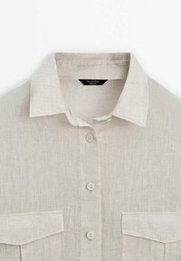 Chemise beige en lin à manches longues avec col pointu, quatre boutons sur le devant et deux poches poitrine à rabats.