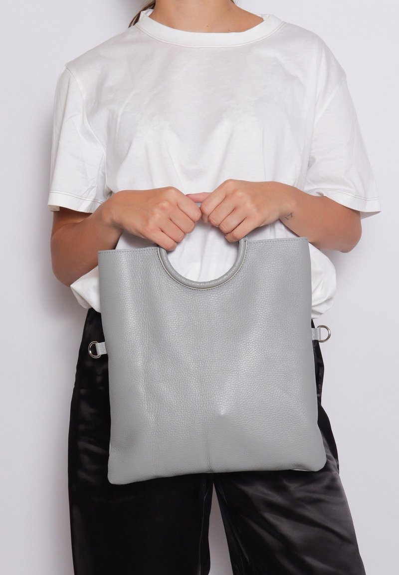 Borsa tote grigia in pelle con manico arrotondato, texture liscia, forma rettangolare e dettagli hardware laterali, tenuta da una persona in camicia bianca.