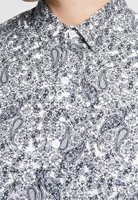 Chemise blanche à boutons avec un motif floral et paisley noir. Le tissu semble lisse avec un col structuré.