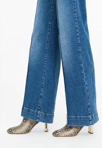 Botas de tobillo de piel de serpiente con punta afilada y tacón bloque dorado, combinadas con pantalones de denim azul de pierna ancha con un dobladillo deshilachado.