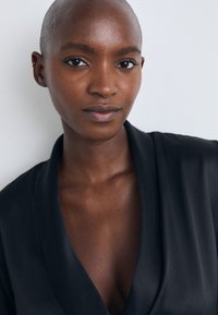 Bald person iklädd en djup V-ringad svart kappa, med slät textur och subtil lyster, har en naturlig hudton och klar hy.