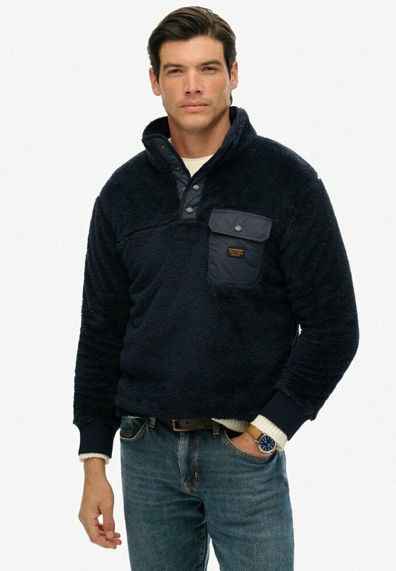 Marine fleece pullover med høj krave, trykknapper og brystlomme. Har ribbede manchetter og krop samt en blød, struktureret overflade.
