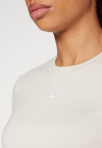 Jasnobeżowa koszulka z okrągłym dekoltem. Na piersi znajduje się biały logo Nike w kształcie łyżwy. Materiał wydaje się gładki i miękki.