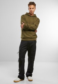 Felpa verde oliva con cappuccio e lacci, abbinata a pantaloni cargo neri con più tasche e dettagli con zip, indossata con sneakers bianche.