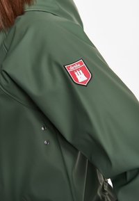 Groene waterdichte jas met een rood-wit schildpatch met een torenontwerp op de linker mouw en zilveren drukknoopsluitingen.