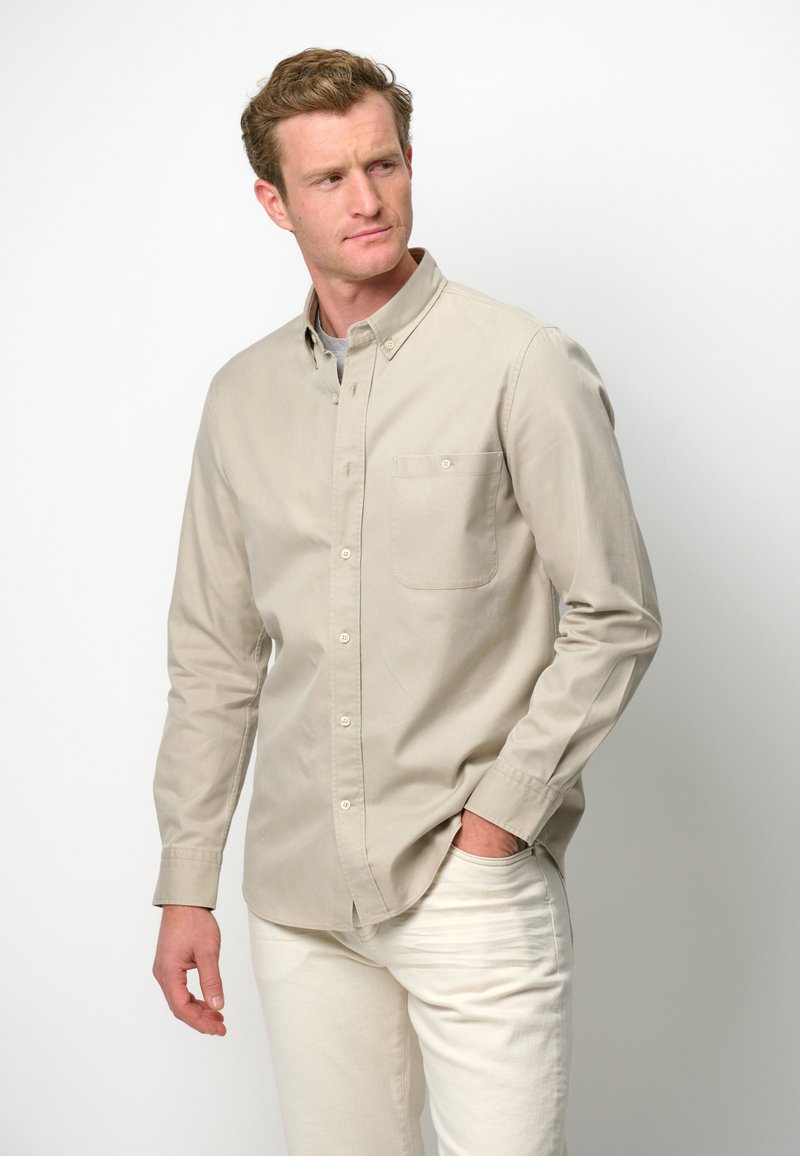 Camicia beige a maniche lunghe con bottoni e tasca sul petto, realizzata in un tessuto morbido. Abbinata a pantaloni chiari, con un fit rilassato.