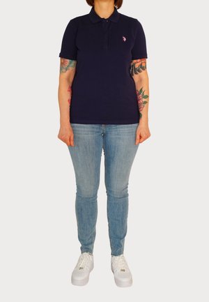 Polo bleu marine avec un logo rose, porté avec un jean bleu clair et des baskets blanches. Manches courtes, col classique et coupe légèrement ajustée.