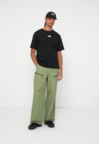 Tricou negru cu logo alb, pantaloni cargo verzi cu buzunare cu fermoar și adidași negri. Modelul poartă o șapcă asortată. Fundal simplu.