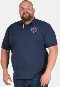 Polo en marine avec col, manches courtes et un patch logo sur la poitrine. Le tissu semble lisse avec une coupe classique.