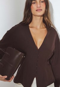 Brūns blūze ar dziļu V veida kaklu un pārmērīgiem piedurknēm, ar pogu aizdari. Modelis tur tumši brūnu ādas rokassomu ar divām rokturiem.