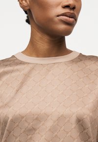 Κοντινό πλάνο ενός ατόμου που φοράει μπεζ πουκάμισο με σχέδιο και στρογγυλή λαιμόκοψη, μπροστά σε απλό φόντο.