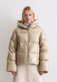 Beżowa pikowana kurtka z kapturem, z zamkiem błyskawicznym z przodu, puchową pikowaną konstrukcją i szerokimi rękawami. Noszona na jasnym dzianinowym golfie.