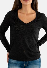 Donna che indossa una maglietta nera a maniche lunghe con stampa leopardata e scollatura a V, e jeans blu, con le mani sui fianchi.