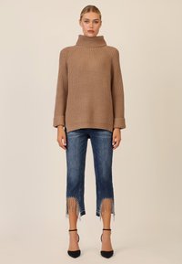 Maglione lavorato a maglia beige con colletto alto, abbinato a jeans blu cropped con frange e perline all'orlo, completato da tacchi neri.