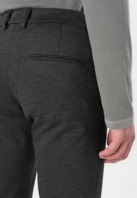 Dunkelgraue Hose mit tailliertem Schnitt, ausgestattet mit einer einzelnen Gesäßtasche und dezenter Textur. Glattes Material mit leichtem Glanz.