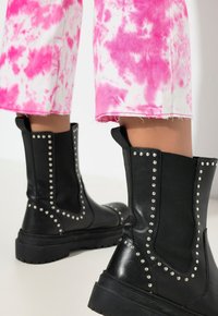 Botas negras hasta el tobillo con tachuelas plateadas a lo largo de las costuras, hechas de cuero y tela. Combinadas con pantalones cortos de teñido anudado rosa.