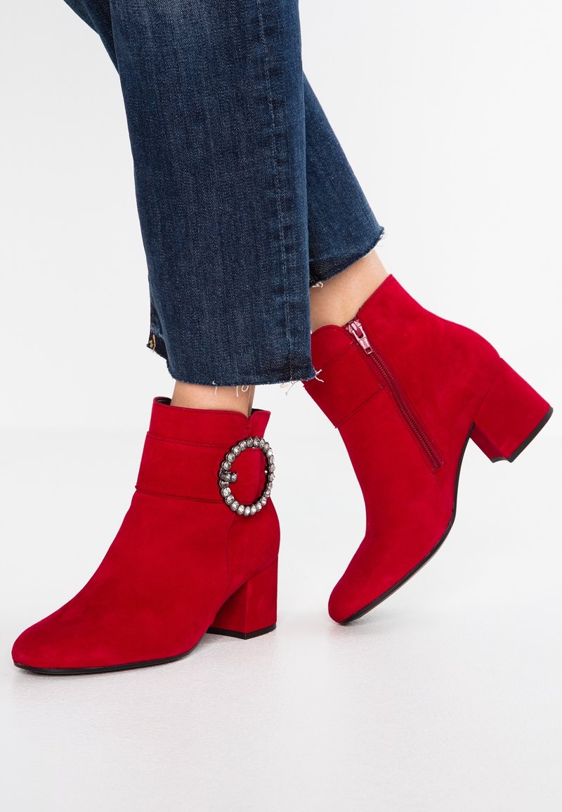 Femme portant des bottines en daim rouge vif à talons blocs, avec une boucle en strass, une fermeture éclair latérale, assorties à un jean court bleu foncé.
