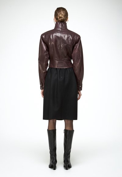 Femme aux cheveux attachés en chignon portant une veste courte en cuir bordeaux, une jupe noire au genou, des collants transparents et des bottes noires montantes, vue de dos.