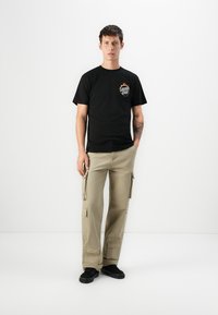 Sort bomulds T-shirt med et frontgrafik; parret med beige cargo bukser, der har flere lommer og en afslappet pasform. Sorte sneakers.